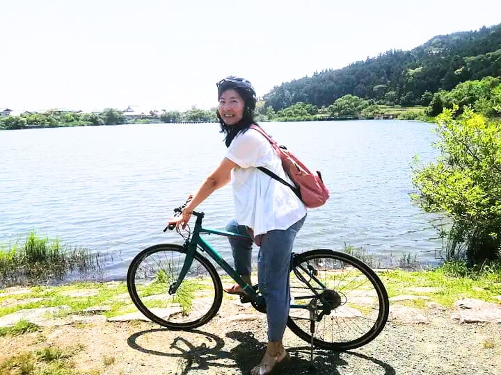 Woman wearing a helmet and casual attire, standing beside a bicycle near a lake.