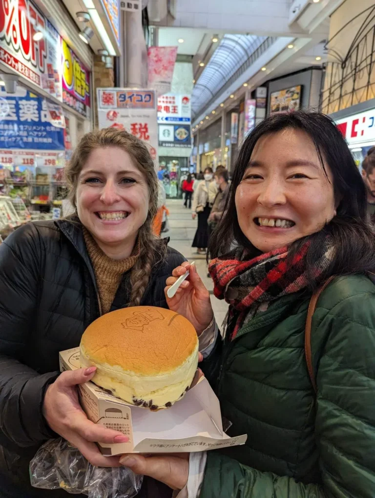 Two women smiling while holding a large cheesecake in a busy street market in Japan.