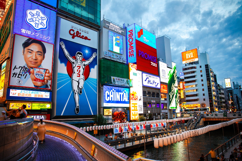 Blogs 4 Osaka Tourist Attractions, Bright neon signs and advertisements illuminating a cityscape at dusk with a river in the foreground.