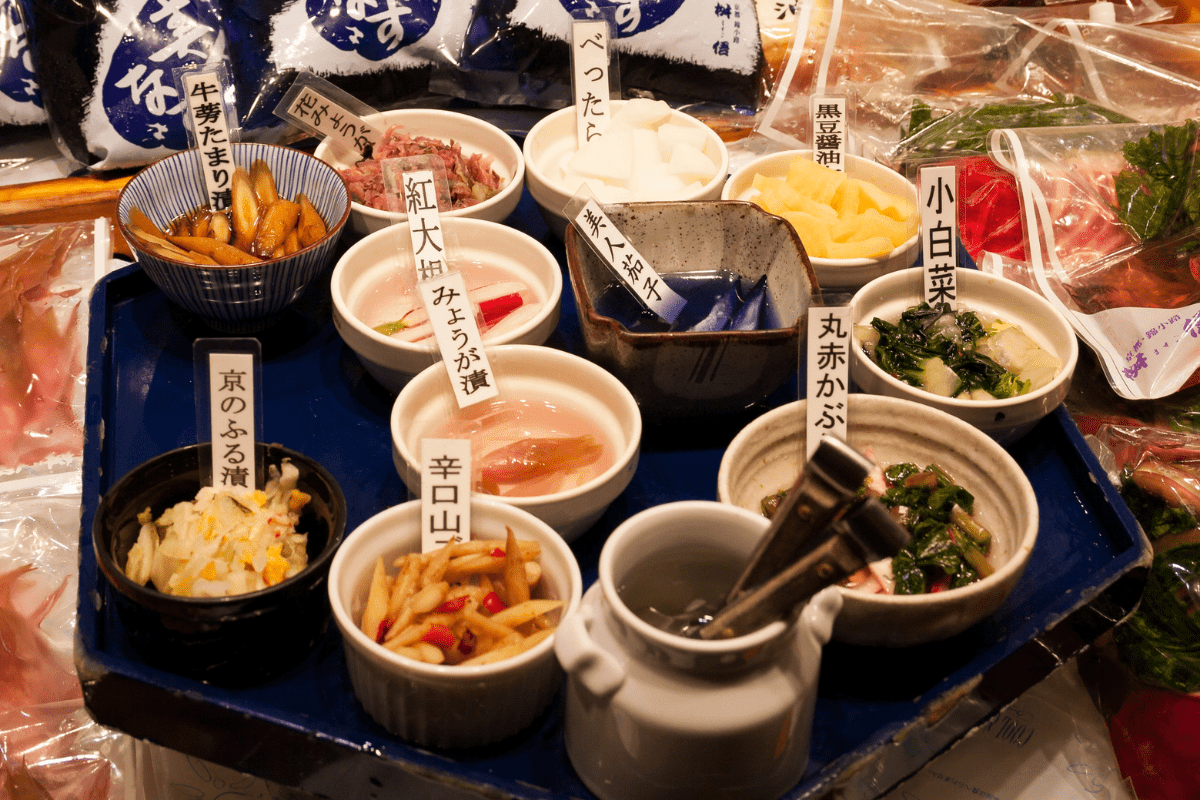 15 Irresistible Traditional Kyoto Foods You Need to Try 4 A tray displays assorted Japanese pickled vegetablesāa showcase of regional specialties and traditional foodāin small bowls, each labeled in Japanese. The colorful pickles vary in type and color, surrounded by packaged goods and fresh vegetables.