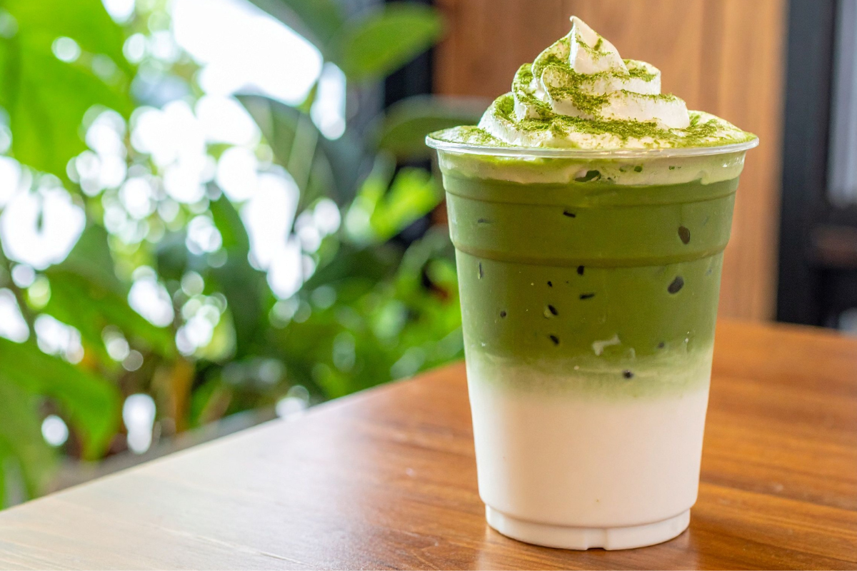 15 Irresistible Traditional Kyoto Foods You Need to Try 13 A plastic cup of iced matcha latte with white milk on the bottom, green matcha on top, and whipped cream with matcha powderāreminiscent of Japanās street food cultureāsits on a wooden table with blurred green plants in the background.
