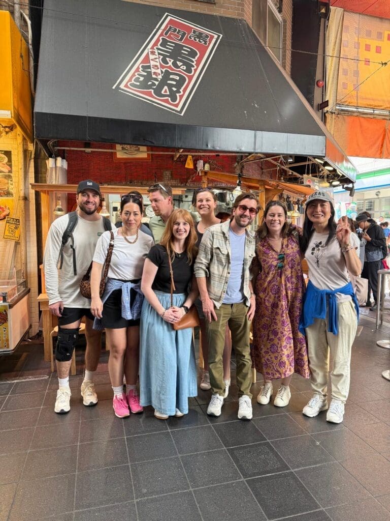 Group of eight people posing in front of a food stall with a black awning and signage in a busy street.