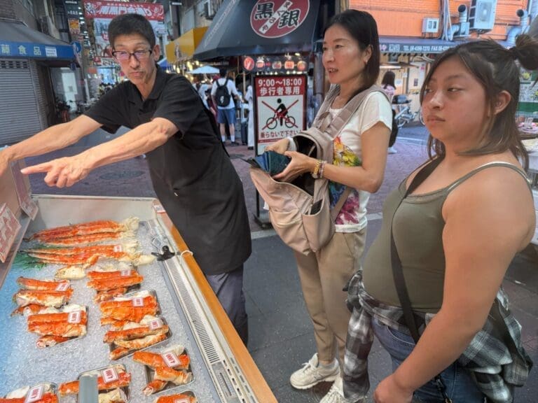 A vendor pointing at a display of seafood while two women observe in a market setting.