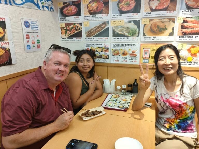 Three individuals seated at a table in a restaurant, enjoying food and smiling at the camera.
