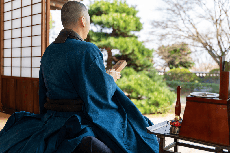 Blogs 25 Koyasan from Osaka, A monk wearing traditional blue robes seated with a wooden object in hand, facing a garden view.