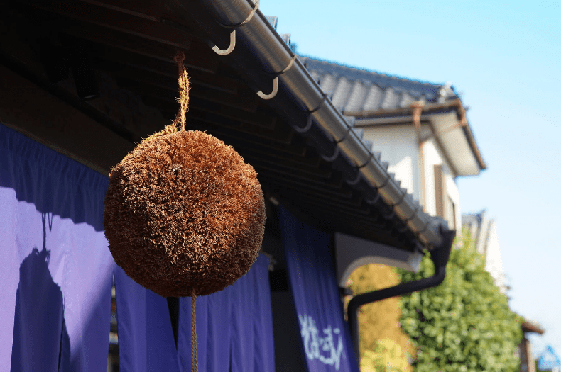 Blogs 17 Kobe Sake Brewery, A round natural fiber ball hanging from a roof eave, with a blue sky in the background.