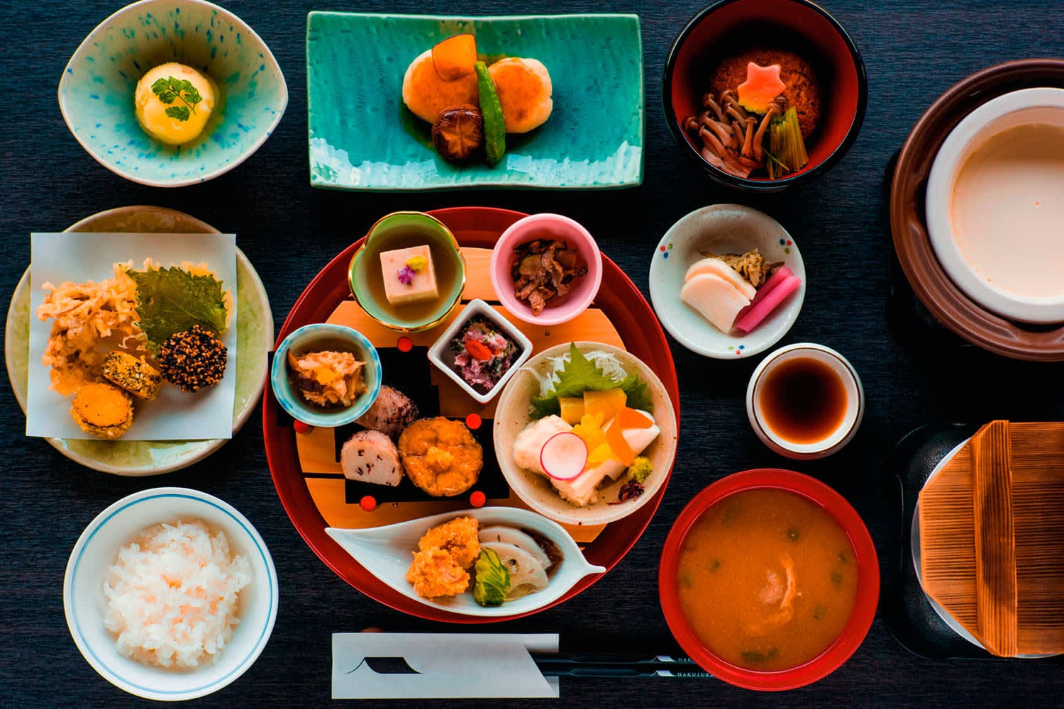 A colorful Japanese kaiseki meal, showcasing Traditional Food in Kyoto, is arranged on a table with rice, soup, pickles, vegetables, tofu, and regional specialties—each beautifully presented in bowls and plates. Chopsticks and a napkin are placed in front.