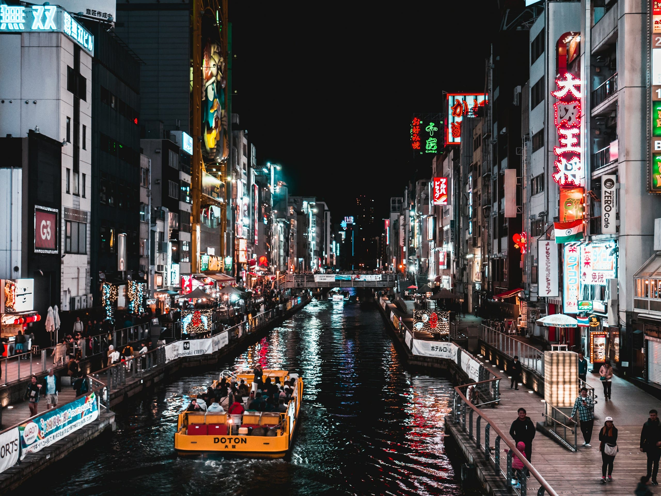 Home 14 A brightly lit urban canal at night showcases the vibrant nightlife in Osaka, with a yellow sightseeing boat, neon signs on tall buildings, and people strolling along both sides of the water.