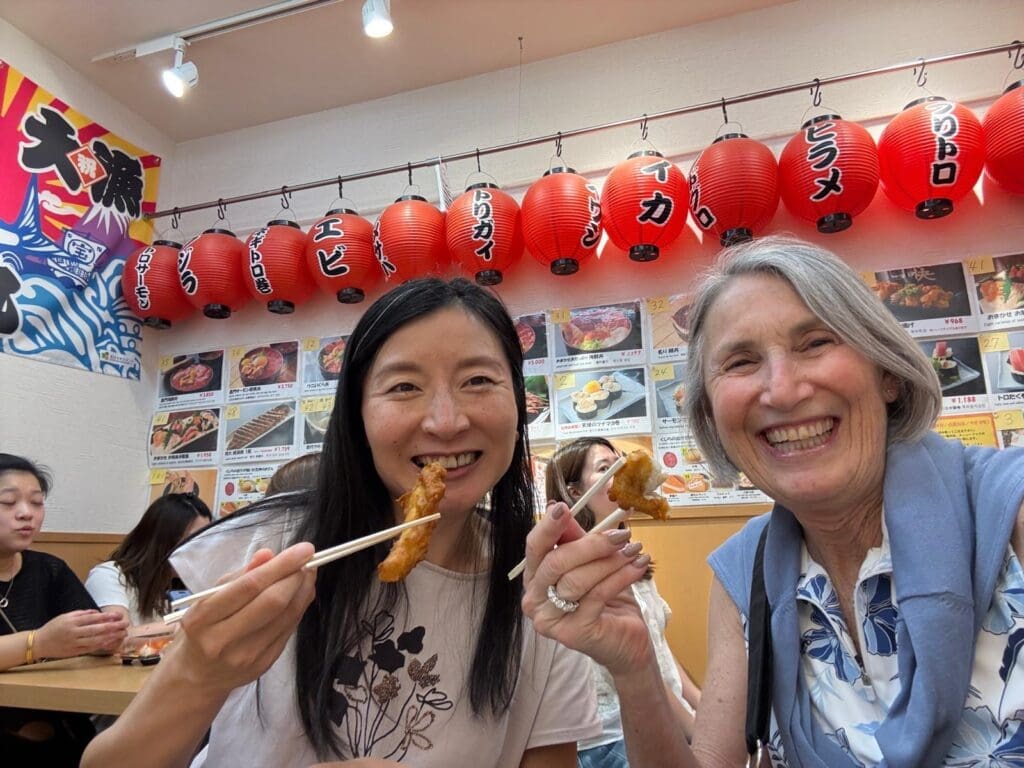 Two women smiling while holding skewers of food with chopsticks in a restaurant with red lanterns.