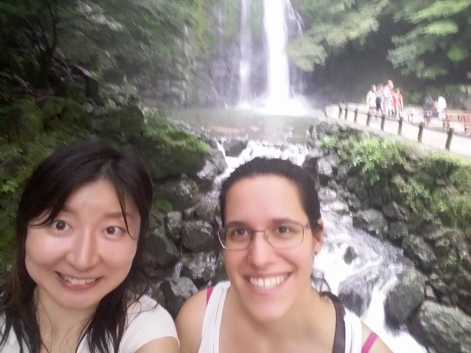 Two women smiling in front of a waterfall surrounded by greenery and rocks in Minoh Park.