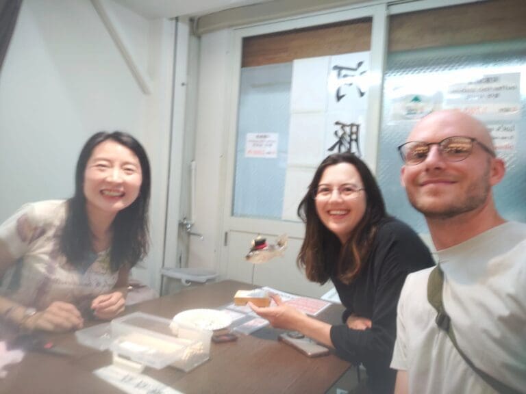 Three individuals smiling at a table with various items, including a small model, in a casual indoor setting.