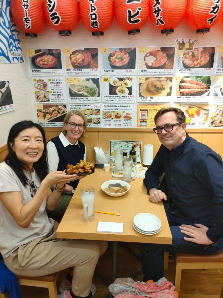 Three individuals seated at a table in a restaurant, with one holding a plate of food and a menu in the background.