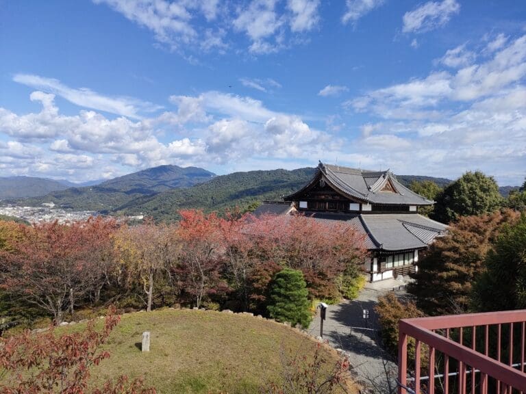 Traditional Japanese temple surrounded by autumn foliage and mountains under a blue sky with clouds.